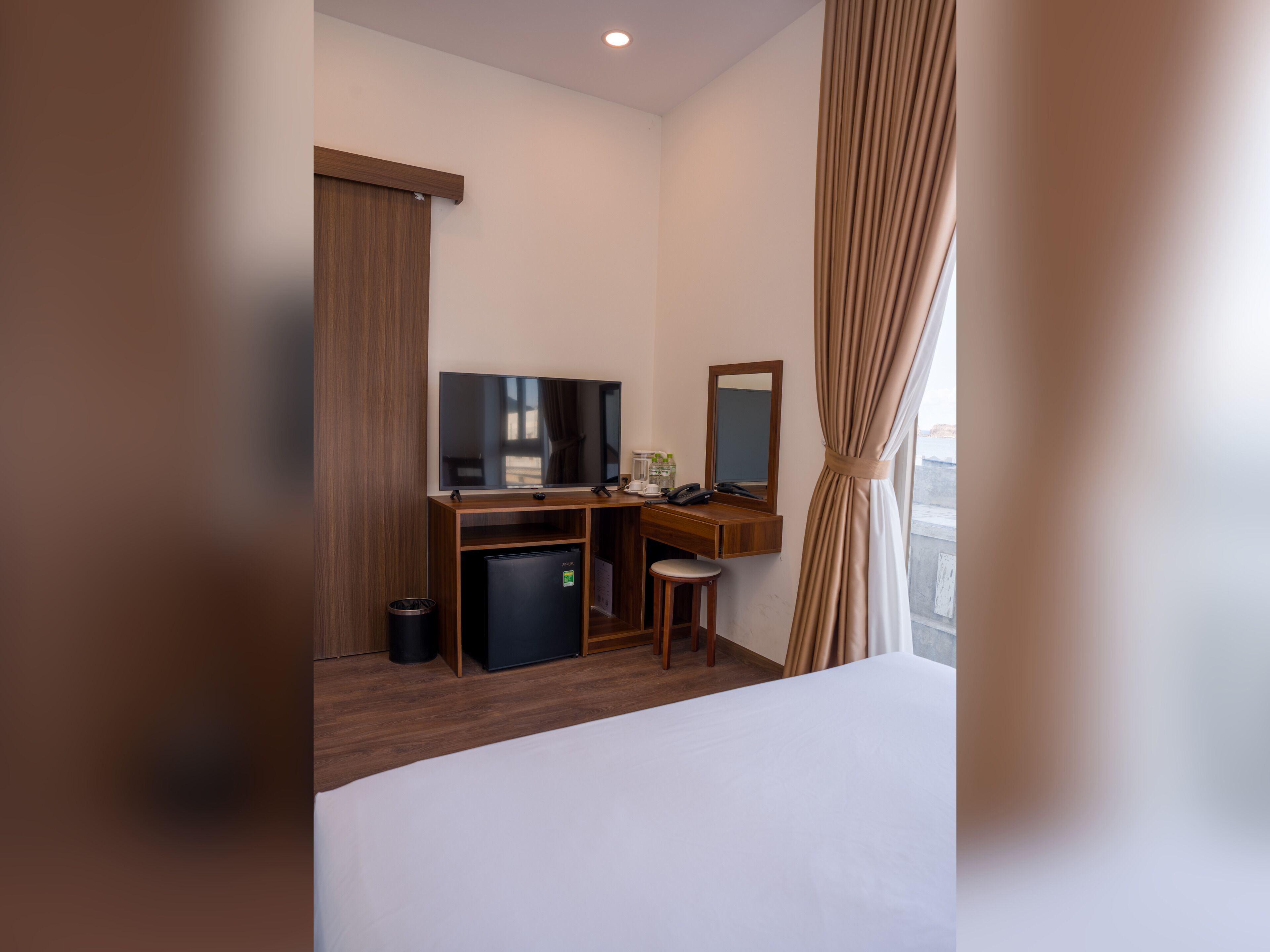 Une chambre d’hôtel équipée d’un lit, d’un bureau avec un ordinateur, d’une télévision, d’un petit réfrigérateur et d’une fenêtre avec des rideaux.