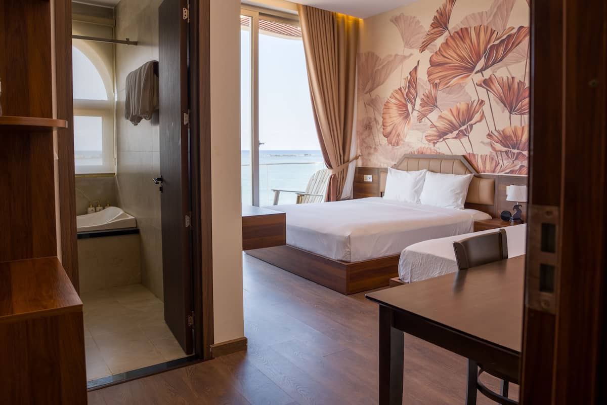 A hotel room with a bed, bedside table, a desk, a chair, a bathroom with a shower, and a view of the sea.