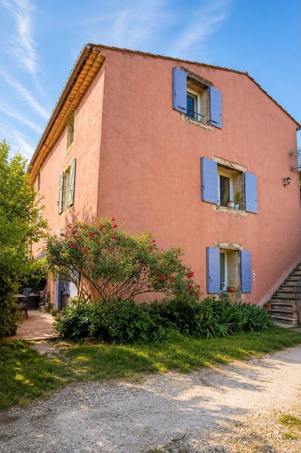 A pink building with blue shutters, a gravel path, and a staircase leading to the entrance.