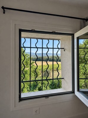 A window with a metal grid, a white wall, and a view of greenery and distant hills.
