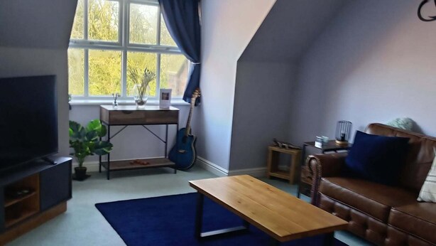 A living room with a TV, a wooden coffee table, a brown sofa, a blue rug, a guitar, and a window with curtains.