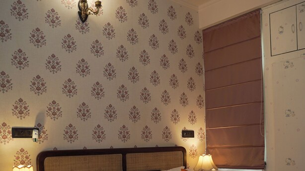 A bedroom with a bed, two bedside lamps, patterned wallpaper, a window with blinds, and a nightstand with a lamp. - Parkside Haven 3BHK By Avinal (Jaipur)