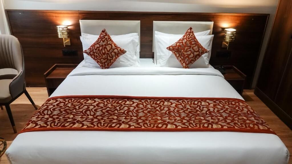 A hotel room with a large bed, two nightstands, a chair, and a patterned throw blanket.