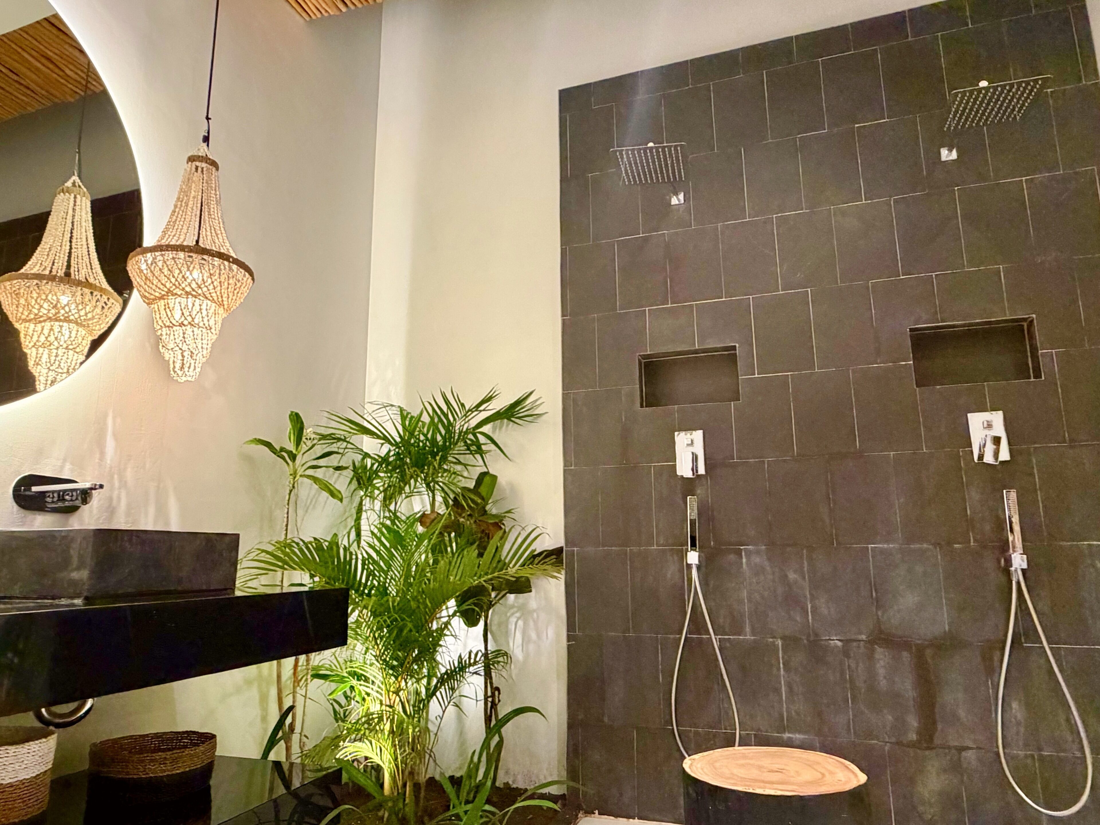 A modern bathroom with a black sink, two hanging lights, a shower area with a control panel, and a potted plant.