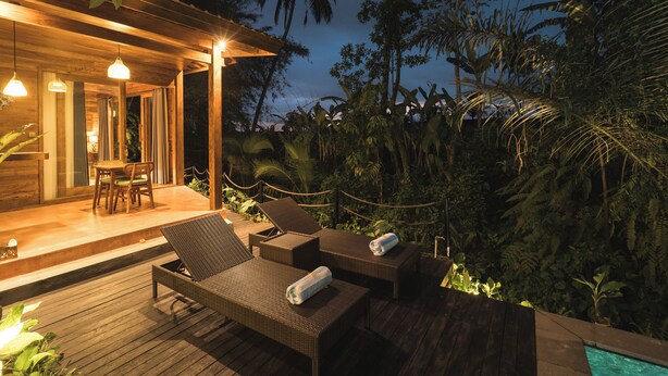 Una terraza de madera con sillones de descanso y una pequeña mesa, rodeada de exuberante vegetación tropical.