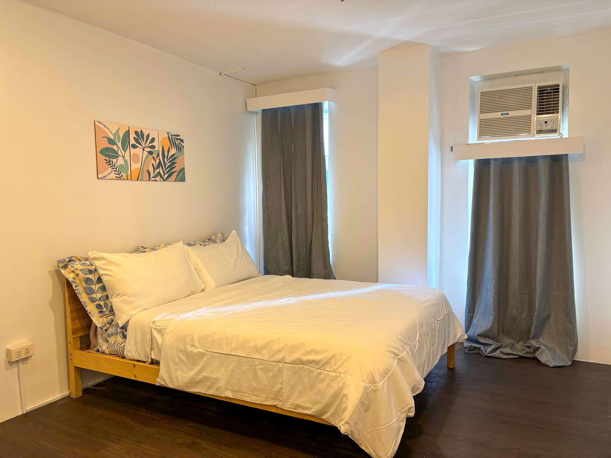 Una cama individual con estructura de madera, ropa de cama blanca y un aire acondicionado de pared.