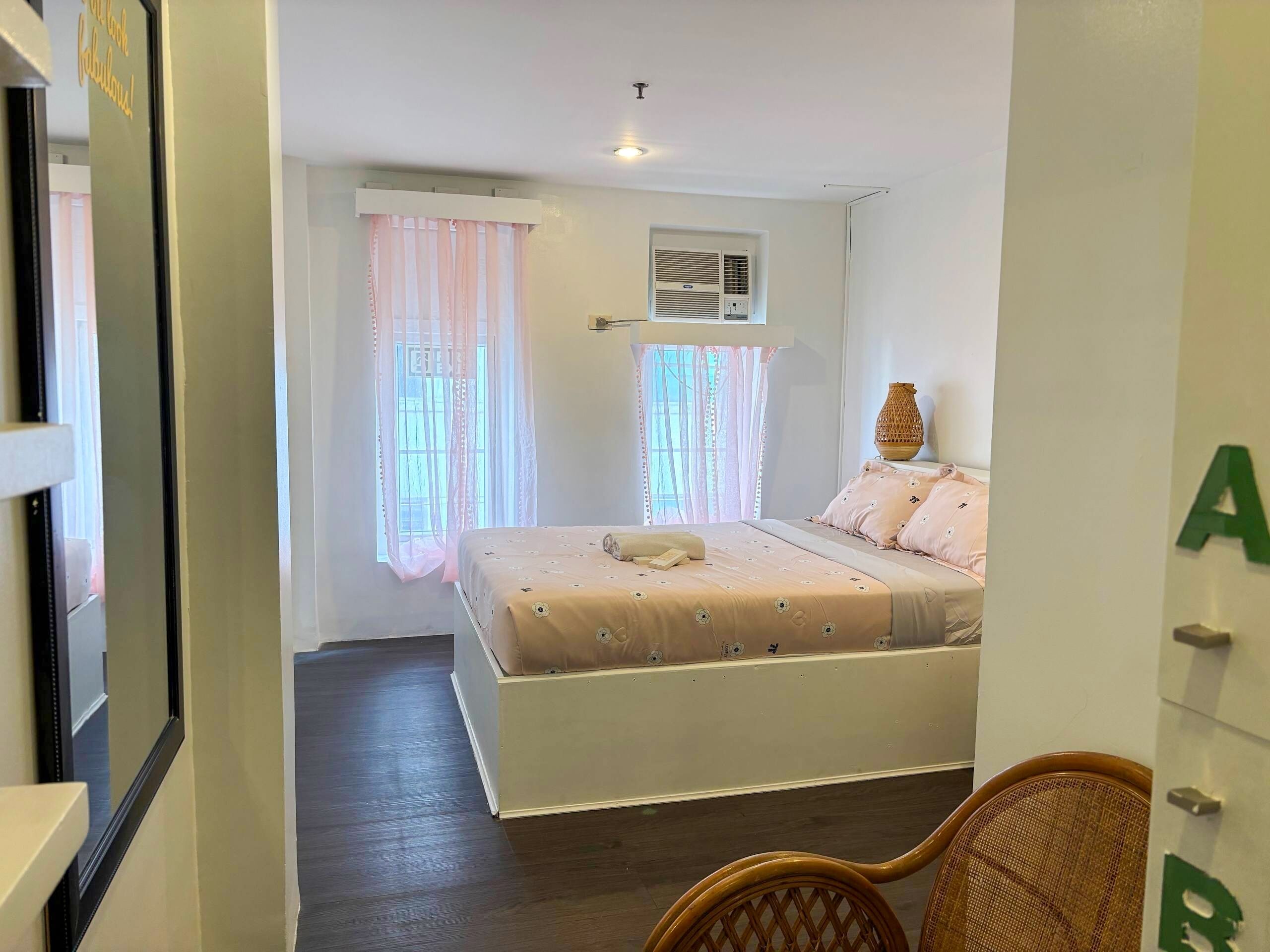 A bedroom with a bed, two windows with curtains, an air conditioning unit, and a wicker chair.