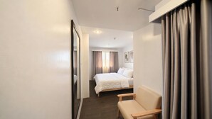 A hotel room with a bed, a chair, a mirror, and a window with curtains. - V Saigon Suites by SMS Hospitality (Makati)