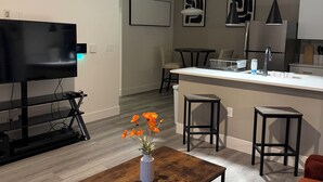 Un salon moderne doté d’un téléviseur à écran plat, d’une table basse ornée d’un vase de fleurs, d’un coin repas avec un îlot de cuisine et d’un bar avec des tabourets.