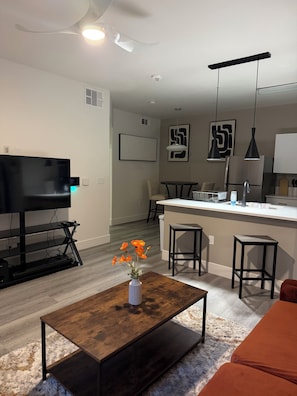 A modern living room with a flat-screen TV, a coffee table with a vase of flowers, a dining area with a kitchen island, and a bar area with stools. - Cozy 1-bedroom apartment in Spring Valley, Las Vegas 10 Minutes from Airport (Las Vegas)