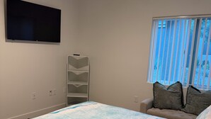 Une chambre à coucher avec un lit, un téléviseur fixé au mur, une fenêtre avec des stores et une table de chevet avec une lampe.