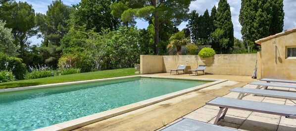 A rectangular swimming pool with clear water, surrounded by a low wall and a small garden area with a few trees and shrubs.