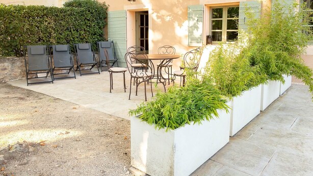 An outdoor seating area with a table and chairs, a hedge, and a building with shutters. - Luberon Cottage at Château Juvenal (Saint-Hippolyte-le-Graveyron)