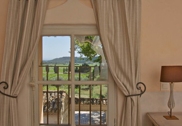 A room with a window overlooking a rural landscape, curtains, a lamp, and a stone wall.