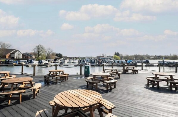 Une terrasse en bois avec des tables de pique-nique, donnant sur une marina où sont amarrés des bateaux.
