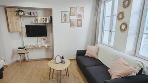 A modern living room with a black sofa, a small wooden table, a TV mounted on a wooden cabinet, and a window with blinds. - Cosy studio at the foot of the ramparts (Boulogne-sur-Mer)