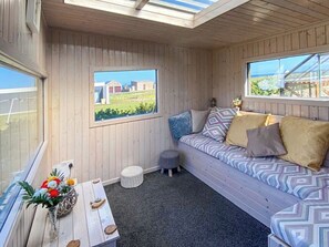 Ein gemütliches Zimmer mit Holzbalkendecke, einem Fenster mit Blick auf ein grünes Feld und Gebäude, einem gemusterten Sofa mit Kissen, einem kleinen runden Hocker und einem Holztisch mit Blumenvase.