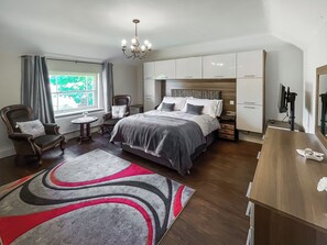 Une chambre à coucher comprenant un lit, une chaise, un bureau, un fauteuil avec un coussin, une petite table, un lustre et une fenêtre avec des rideaux.