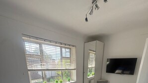 Makuuhuoneessa on sänky, televisio, patteri, ikkuna, jossa on sälekaihtimet, ja tuoli.