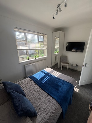 Ein Schlafzimmer mit Bett, Fernseher, Heizkörper, einem Fenster mit Jalousien und einem Sessel.