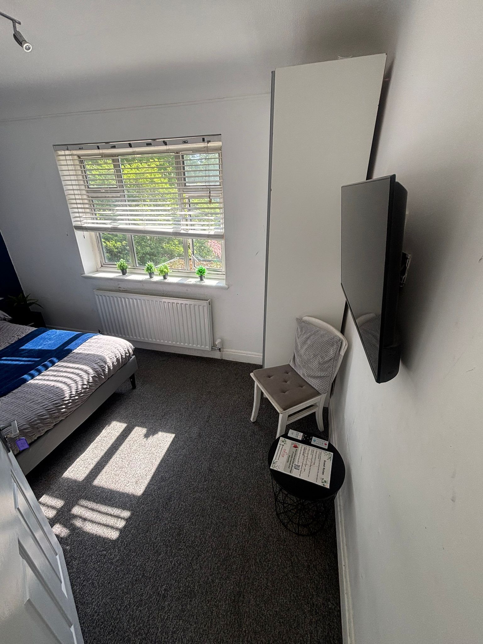 A bedroom with a bed, a window with blinds, a radiator, a chair, and a TV mounted on the wall.