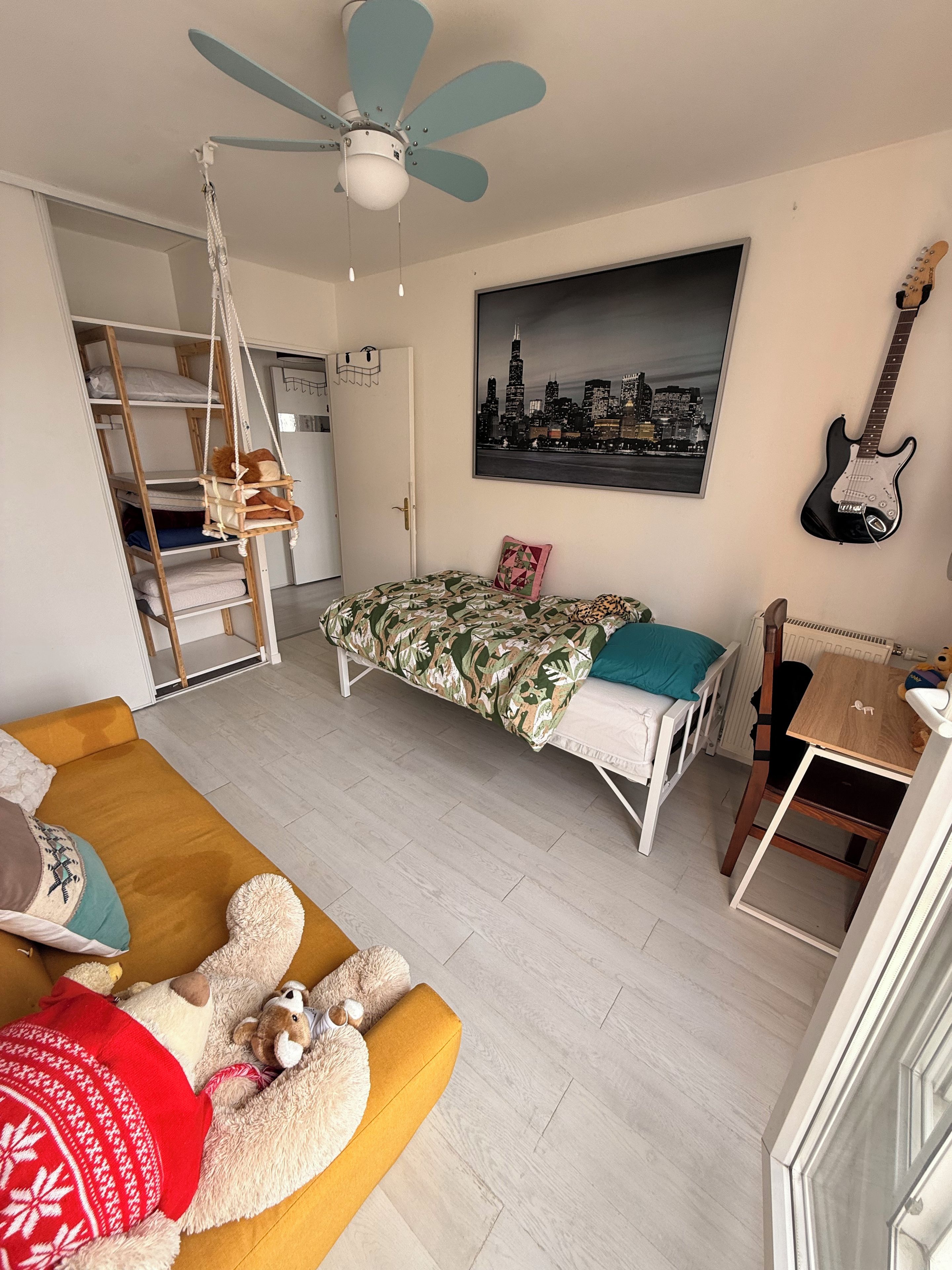 Une chambre à coucher avec un lit, un fauteuil suspendu, une guitare et un tableau représentant un paysage urbain.