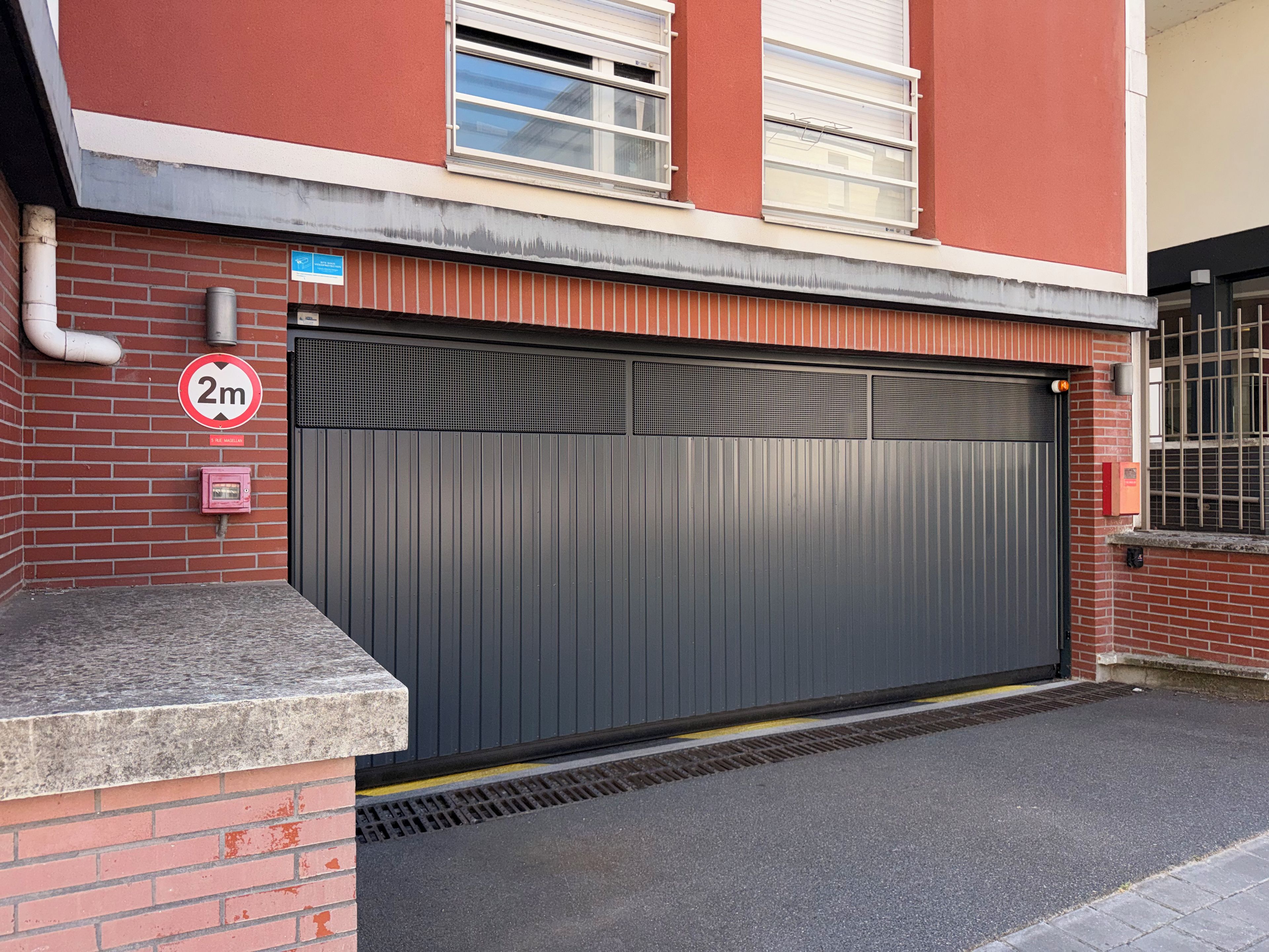 A closed garage door with a 2m height restriction sign, a red brick wall, and a metal vent.
