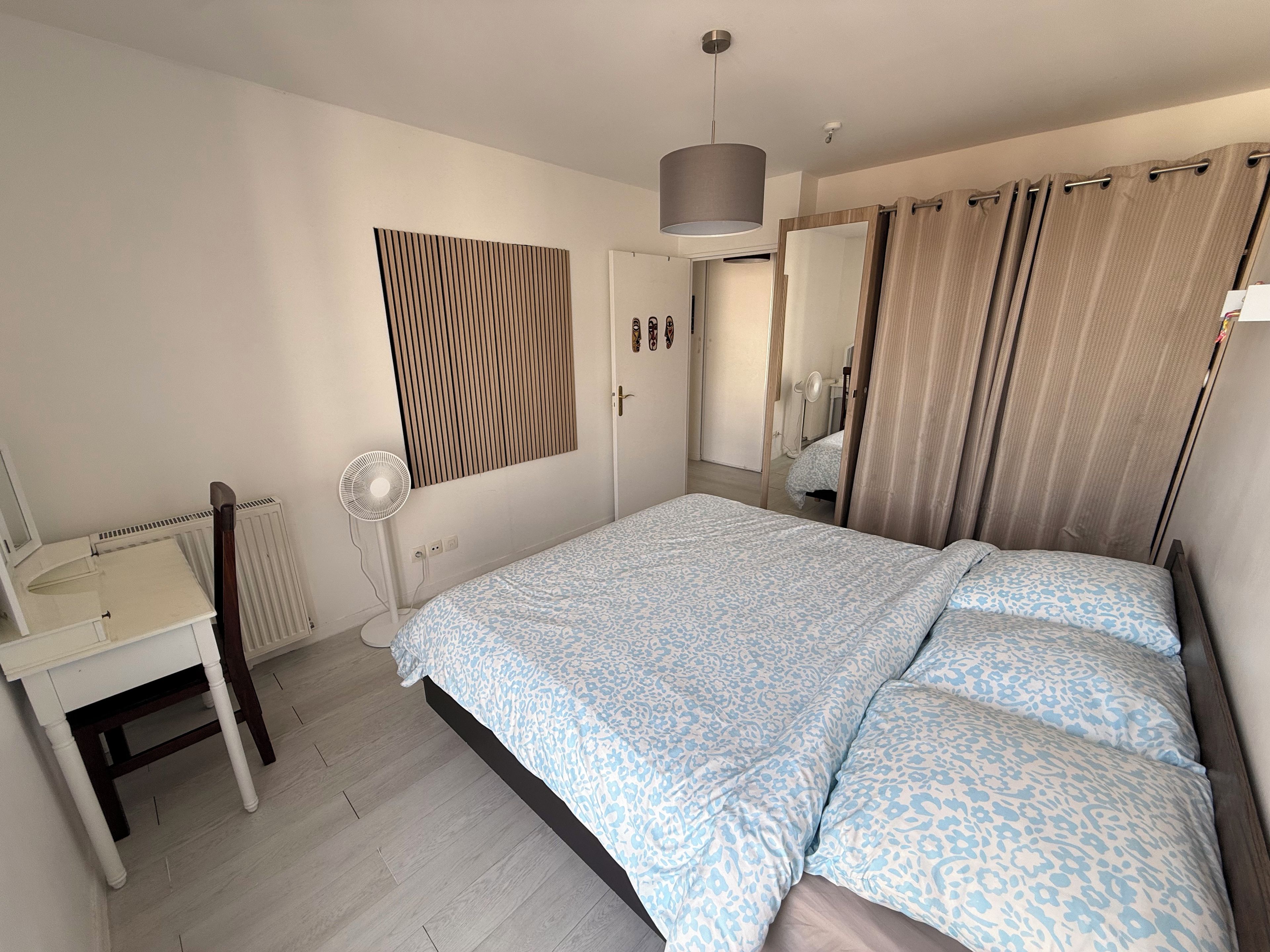 Een slaapkamer met een bed, nachtkastje, spiegel, radiator en een raam met gordijnen.