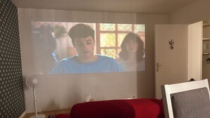 A room with a projector screen, a red sofa, a standing fan, and a door. - Apartment + Parking in Paris, Massy (TGV/RER B/C, Orly Bus) (Massy)