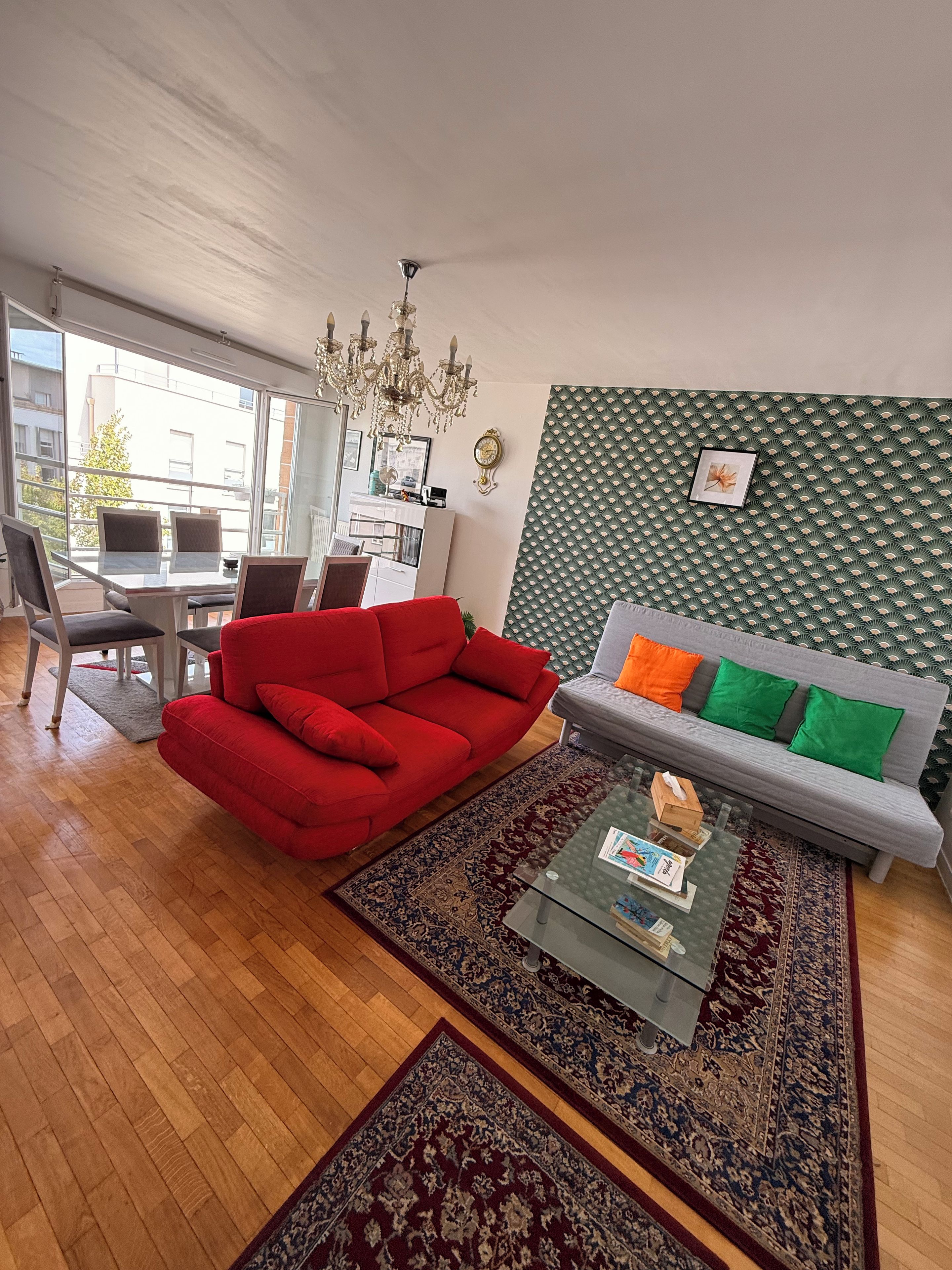 Un salon moderne avec un canapé rouge, un canapé gris agrémenté de coussins verts et orange, une table basse en verre et un mur à motifs.