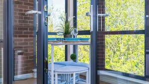 A room with a glass door, a round table with a blue cushion, a potted plant, and a view of trees outside. - Stylish 2-Bed Flat with Parking Near Tube & Heathrow (Uxbridge)
