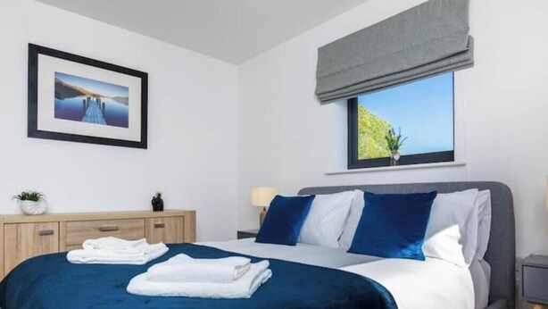 A neatly made bed with a blue bedspread, a grey headboard, and a window with a view of greenery. - Stylish 2-Bed Flat with Parking Near Tube & Heathrow (Uxbridge)