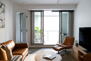 A modern living room with a sofa, armchair, coffee table, TV, and a balcony with a table and chairs. - Premium St Kilda Rd Apt Pool, Gym and Park Access (St Kilda)