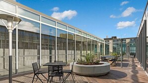 Un espace extérieur aménagé en terrasse, avec des tables et des chaises, un jardinier avec des plantes et une façade de bâtiment en verre.