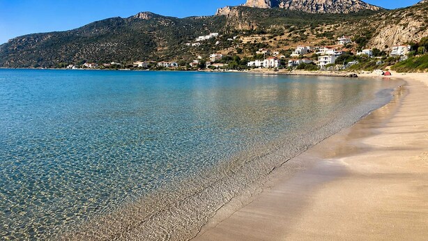 Ein Strand mit klarem Wasser, sandigem Ufer und Gebäuden im Hintergrund.