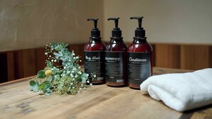 Drie flessen douchegel, shampoo en conditioner op een houten tafel met een witte handdoek en wat groen.