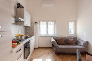 A compact kitchen with a stove, oven, and white cabinets, a sofa with patterned cushions, and a window with curtains. - Via Chiappori Sestri Ponente (Genoa)
