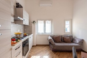 A compact kitchen with a stove, oven, and white cabinets, a sofa with patterned cushions, and a window with curtains.