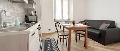 Eine moderne Küche mit Einbauherd, Spüle und Kaffeemaschine. Ein Essbereich mit Tisch und Stühlen sowie ein schwarzes Sofa mit Kissen.