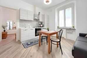 A modern kitchen with white cabinets, a dining table with chairs, and a black sofa in a bright room with large windows. - Via D'andrade Sestri Ponente (Genoa)