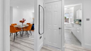 Ein modernes Interieur mit einer Essecke, die einen Glastisch und orangefarbene Stühle sowie eine weiße Tür und eine Küche mit weißen Schränken zeigt.