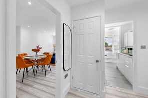 Ein modernes Interieur mit einer Essecke, die einen Glastisch und orangefarbene Stühle sowie eine weiße Tür und eine Küche mit weißen Schränken zeigt.