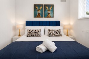 Ein ordentlich bezogenes Bett mit blauem Kopfteil, zwei Kissen mit geometrischen Mustern und zwei an der Wand hängende Hasenbilder.
