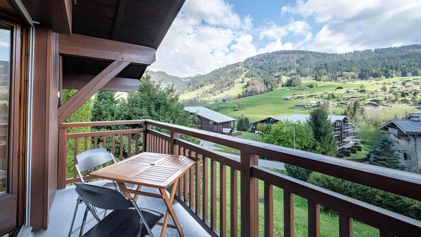 Varanda com mesa e cadeiras, com vista para um vale verde com casas e um lago.