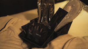 Un letto con una coperta bianca e una borsa nera contenente oggetti con l'etichetta 'VANITY SET' e 'SHOWER'.
