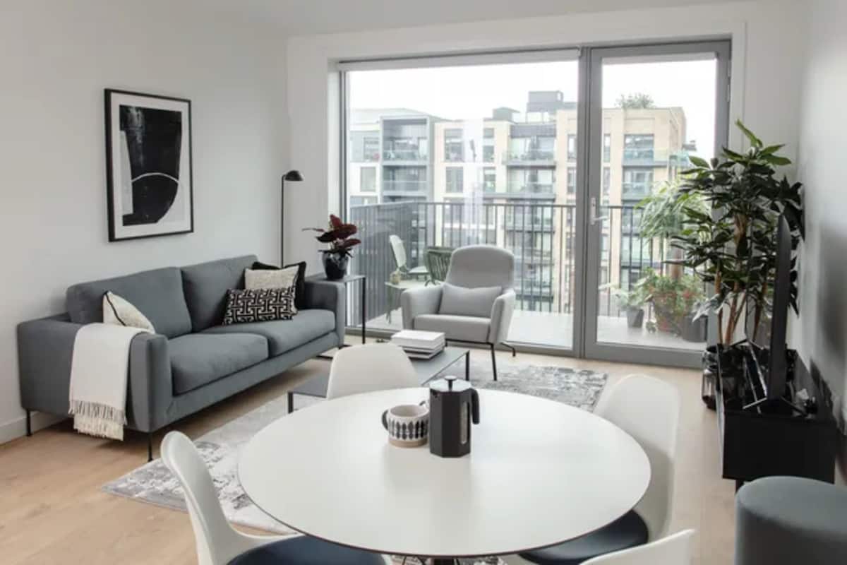 Un moderno salón con un sofá gris, sillas blancas, una mesa redonda y un amplio ventanal con vistas a la ciudad.