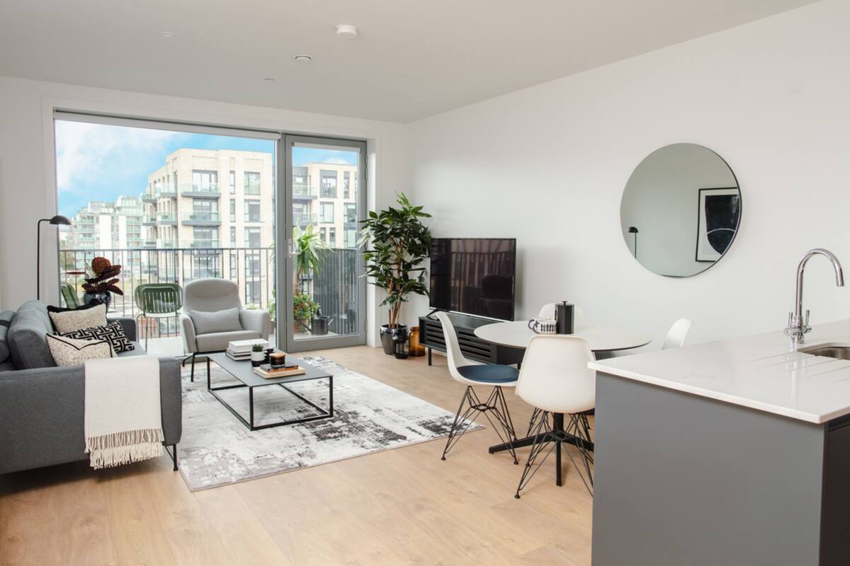 Una sala moderna con sofá, mesa de centro y sillas, un comedor con mesa y sillas, una cocina con lavaplatos y encimera, y grandes ventanas con vistas a la ciudad.