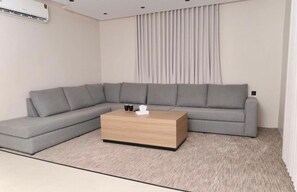 A modern living room with a grey sectional sofa, a wooden coffee table, and a large window with curtains. - Moodar (Madinah)