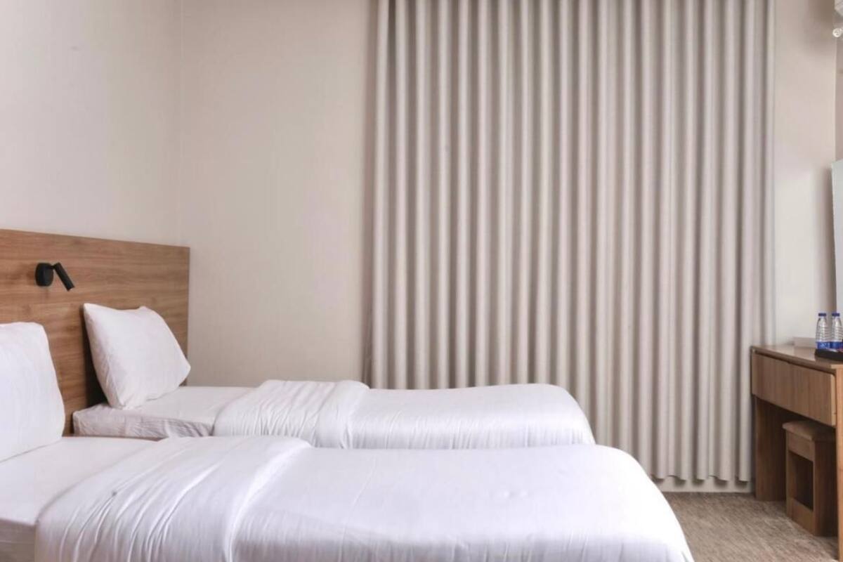 A hotel room with two beds, a wooden headboard, a bedside table with a lamp, and a window with vertical blinds.