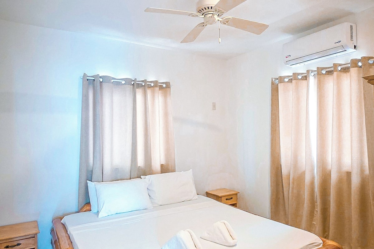 Une chambre à coucher comprenant un lit, un ventilateur de plafond, des rideaux et un climatiseur fixé au mur.